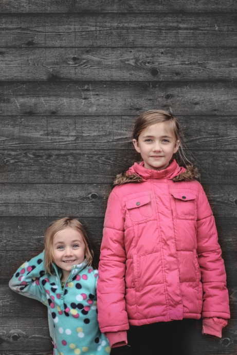 Two girls stand against a wooden wall. Photo by Not So SAHM
